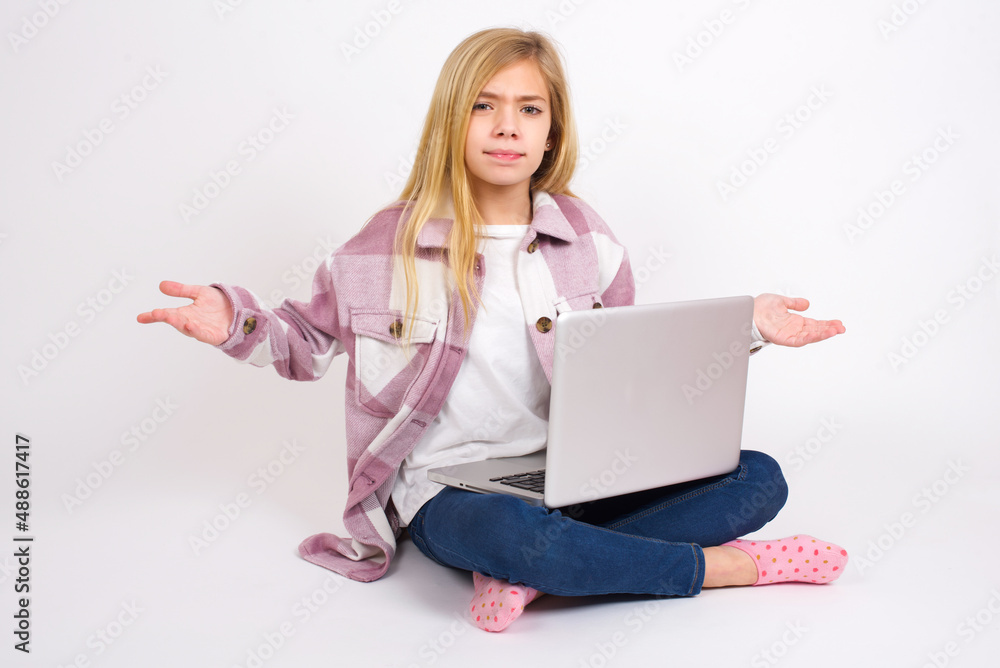 caucasian teen girl sitting with laptop in lotus position on white background looks uncertain shrugs shoulders. caucasian teen girl sitting with laptop in lotus position on white background looks uncertain shrugs shoulders.