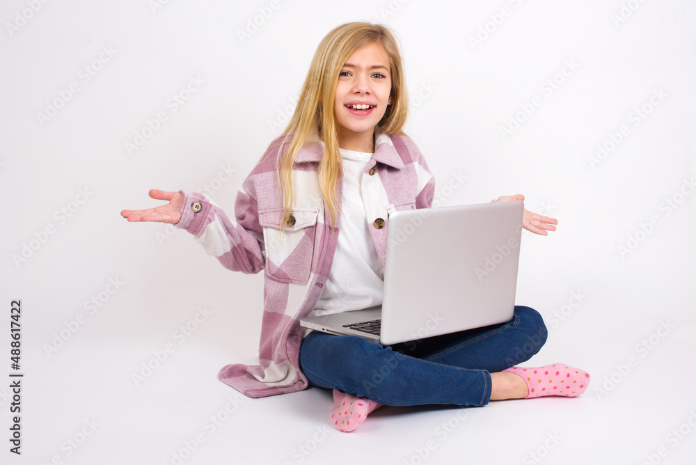 Fototapeta premium So what? Portrait of arrogant caucasian teen girl sitting with laptop in lotus position on white background shrugging hands sideways smiling gasping indifferent, telling something obvious.