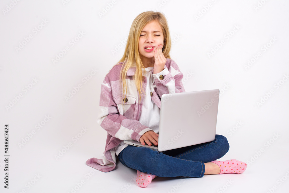 Fototapeta premium Tooth ache concept. caucasian teen girl sitting with laptop in lotus position feeling pain, holding his cheek with hand, suffering from bad toothache, looking at camera with painful expression
