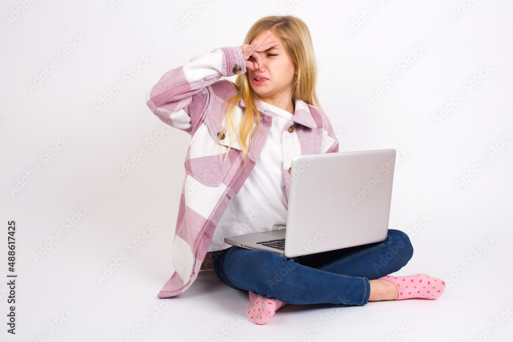 Displeased caucasian teen girl sitting with laptop in lotus position on white background plugs nose as smells something stink and unpleasant, feels aversion, hates disgusting scent. Displeased caucasian teen girl sitting with laptop in lotus position on white background plugs nose as smells something stink and unpleasant, feels aversion, hates disgusting scent.