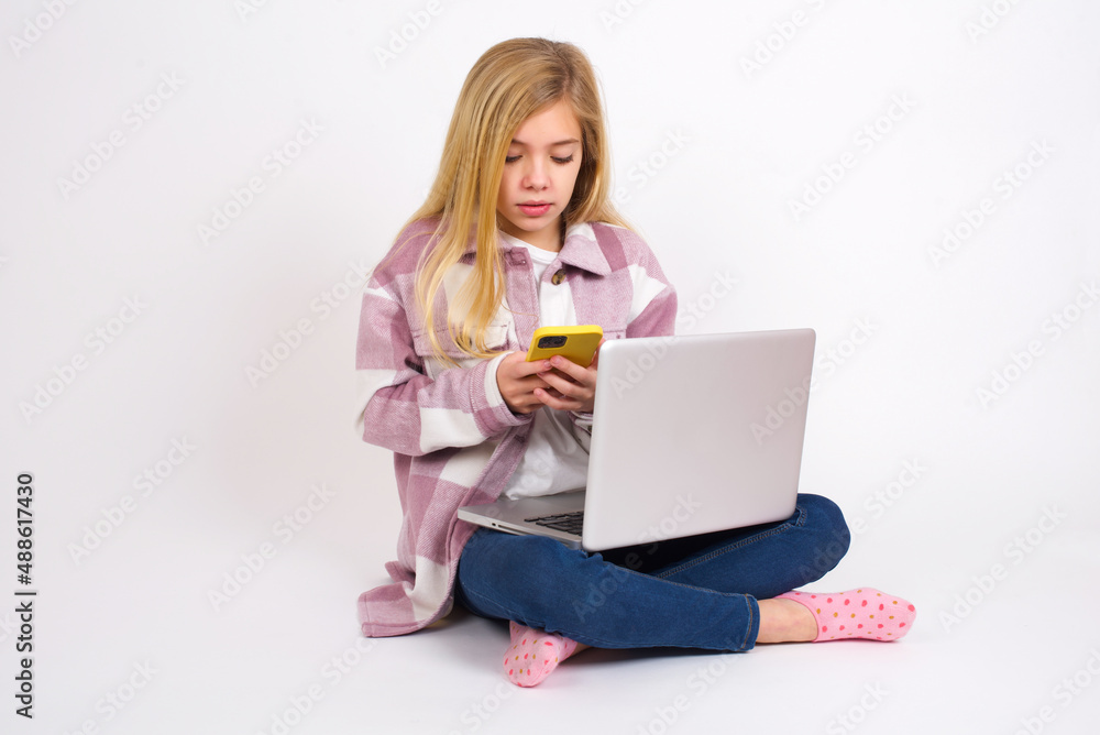 Smiling caucasian teen girl sitting with laptop in lotus position on white using cell phone, messaging, being happy to text with friends, looking at smartphone. Modern technologies and communication. Smiling caucasian teen girl sitting with laptop in lotus position on white using cell phone, messaging, being happy to text with friends, looking at smartphone. Modern technologies and communication.