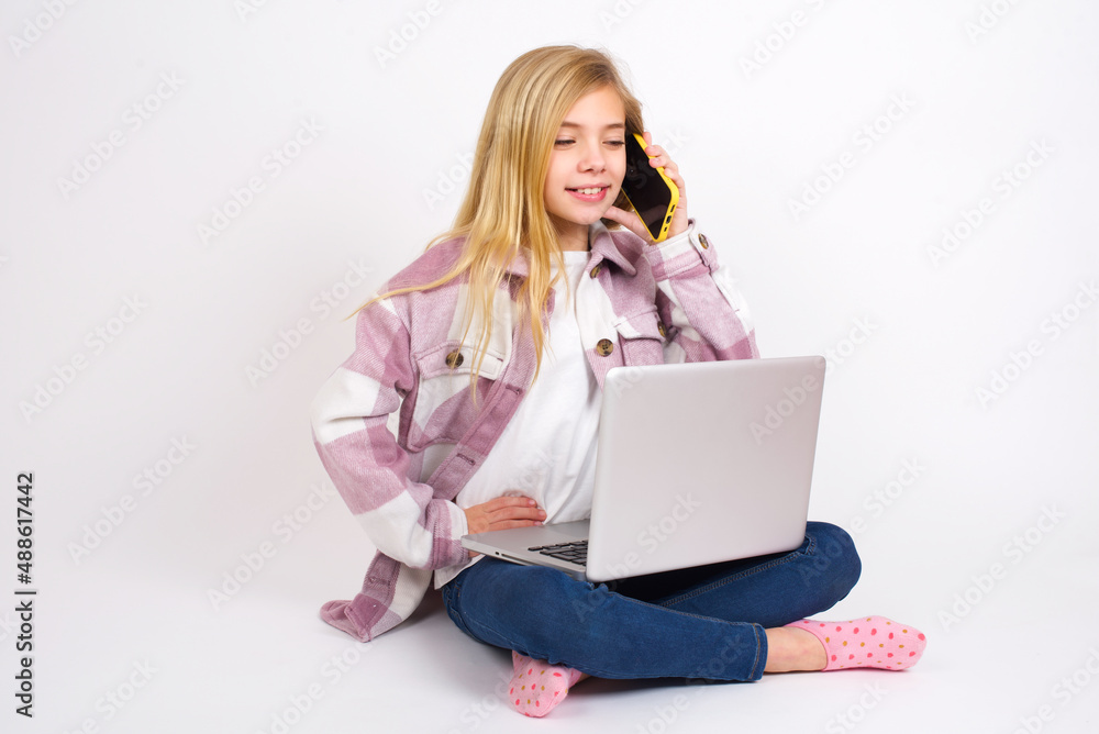 Portrait of successful joyful caucasian teen girl sitting with laptop in lotus position on white background talking on mobile phone with friend. Lifestyle and communication concept Portrait of successful joyful caucasian teen girl sitting with laptop in lotus position on white background talking on mobile phone with friend. Lifestyle and communication concept