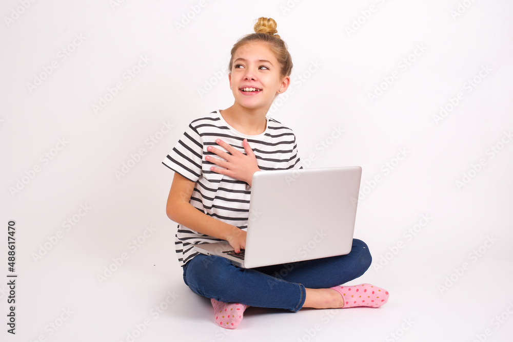 Joyful caucasian teen girl sitting with laptop in lotus position on white background expresses positive emotions recalls something funny keeps hand on chest and giggles happily. Joyful caucasian teen girl sitting with laptop in lotus position on white background expresses positive emotions recalls something funny keeps hand on chest and giggles happily.