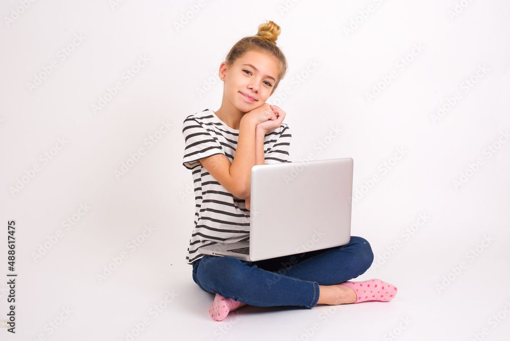 Charming serious caucasian teen girl sitting with laptop in lotus position on white background keeps hands near face smiles tenderly at camera Charming serious caucasian teen girl sitting with laptop in lotus position on white background keeps hands near face smiles tenderly at camera