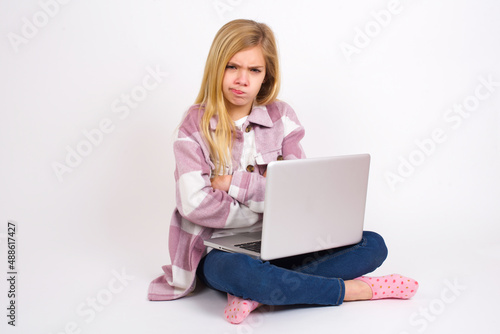 Picture of angry  caucasian teen girl sitting with laptop in lotus position on white background crossing arms. Looking at camera with disappointed expression. © Jihan