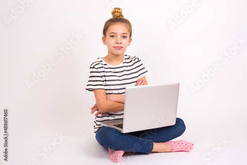 Self confident serious calm caucasian teen girl sitting with laptop in lotus position on white background stands with arms folded. Shows professional vibe stands in assertive pose.