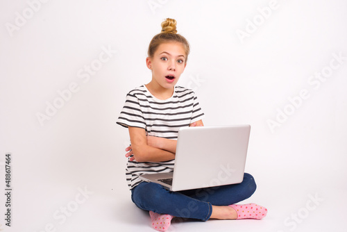 Shocked embarrassed caucasian teen girl sitting with laptop in lotus position on white background keeps mouth widely opened. Hears unbelievable novelty stares in stupor