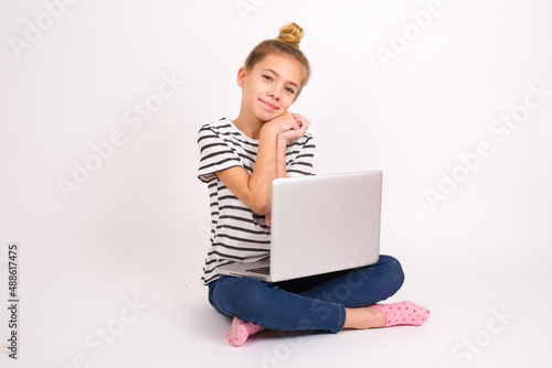 Charming serious caucasian teen girl sitting with laptop in lotus position on white background keeps hands near face smiles tenderly at camera