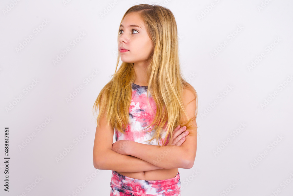 Charming thoughtful little caucasian kid girl wearing sport clothing over white background stands with arms folded concentrated somewhere with pensive expression thinks what to do Charming thoughtful little caucasian kid girl wearing sport clothing over white background stands with arms folded concentrated somewhere with pensive expression thinks what to do