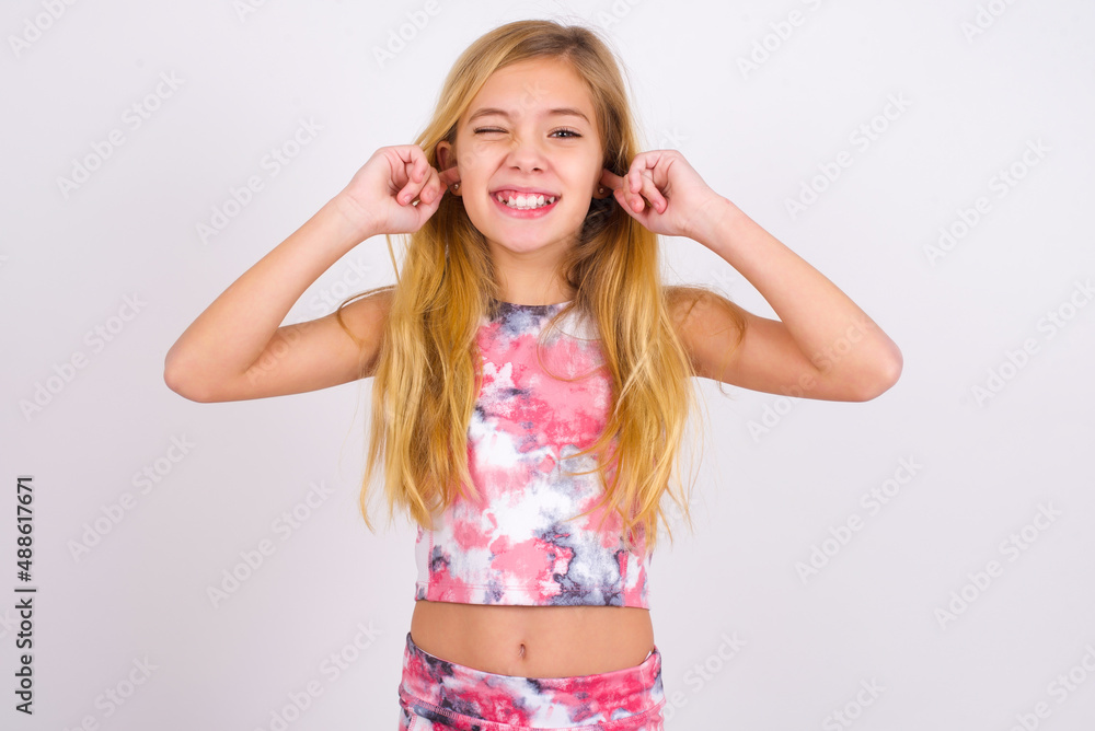 Happy little caucasian kid girl wearing sport clothing over white background ignores loud music and plugs ears with fingers asks to turn off sound Happy little caucasian kid girl wearing sport clothing over white background ignores loud music and plugs ears with fingers asks to turn off sound