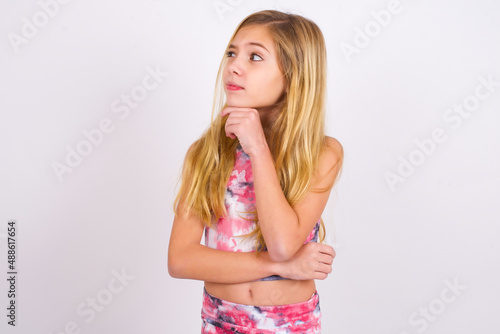 Thoughtful little caucasian kid girl wearing sport clothing over white background holds chin and looks away pensively makes up great plan photo