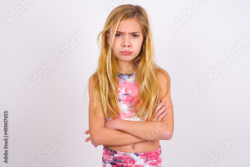 Gloomy dissatisfied little caucasian kid girl wearing sport clothing over white background looks with miserable expression at camera from under forehead, makes unhappy grimace © Roquillo