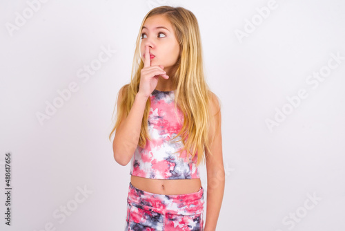 little caucasian kid girl wearing sport clothing over white background silence gesture keeps index finger to lips makes hush sign. Asks not to share secret