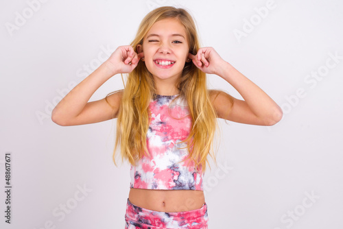 Happy little caucasian kid girl wearing sport clothing over white background ignores loud music and plugs ears with fingers asks to turn off sound