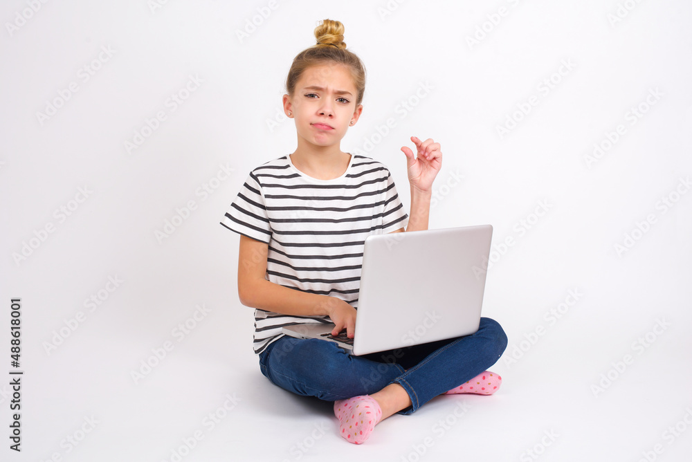 Displeased caucasian teen girl sitting with laptop in lotus position on white background shapes little hand sign demonstrates something not very big. Body language concept. Displeased caucasian teen girl sitting with laptop in lotus position on white background shapes little hand sign demonstrates something not very big. Body language concept.