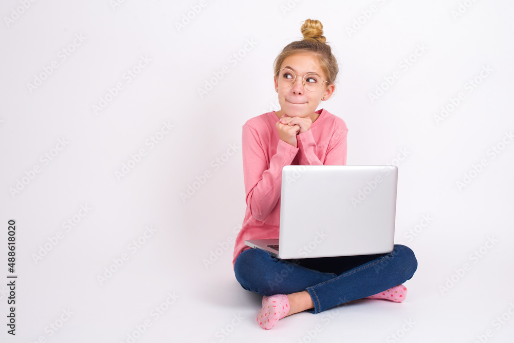 Curious caucasian teen girl sitting with laptop in lotus position on white background keeps hands under chin bites lips and looks with interest aside. Curious caucasian teen girl sitting with laptop in lotus position on white background keeps hands under chin bites lips and looks with interest aside.