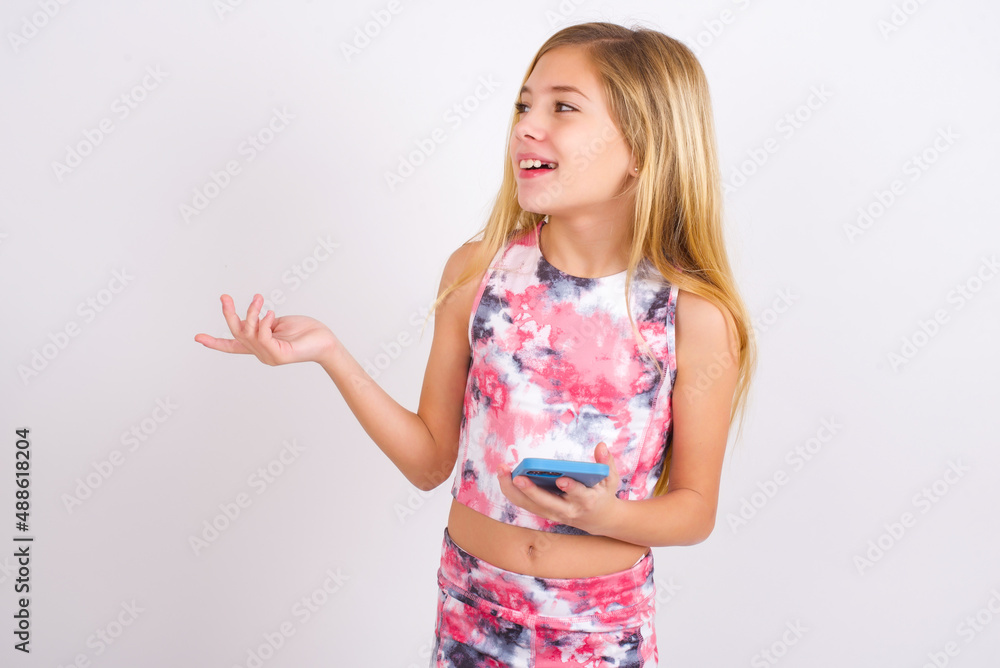Happy pleased little caucasian kid girl wearing sport clothing over white background raises palm and holds cellphone uses high speed internet for text messaging or video calls Happy pleased little caucasian kid girl wearing sport clothing over white background raises palm and holds cellphone uses high speed internet for text messaging or video calls