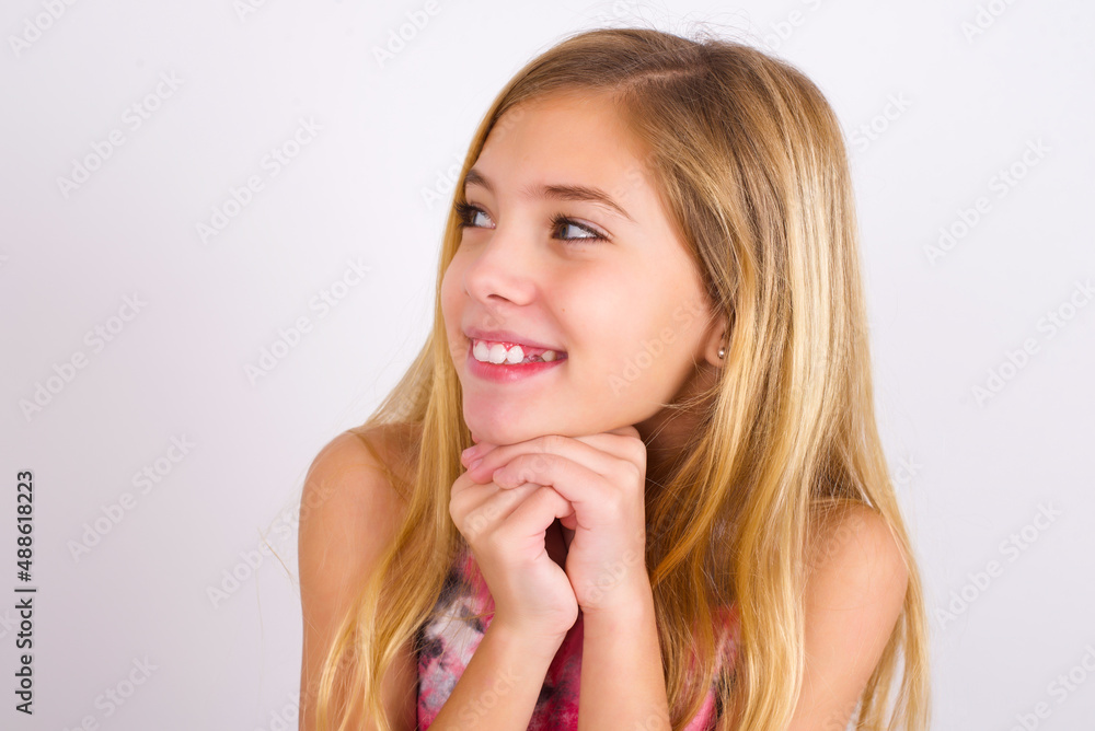 Happy little caucasian kid girl wearing sport clothing over white background anticipates something awesome happen, looks happily aside, keeps hands together near face, has glad expression. Happy little caucasian kid girl wearing sport clothing over white background anticipates something awesome happen, looks happily aside, keeps hands together near face, has glad expression.