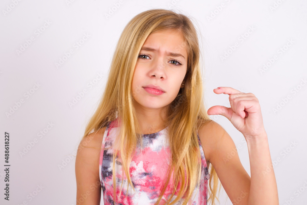 Displeased little caucasian kid girl wearing sport clothing over white background shapes little hand sign demonstrates something not very big. Body language concept. Displeased little caucasian kid girl wearing sport clothing over white background shapes little hand sign demonstrates something not very big. Body language concept.