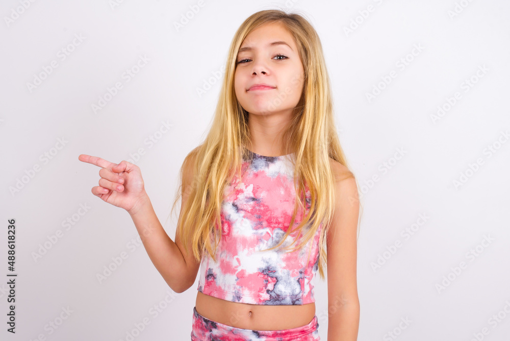 little caucasian kid girl wearing sport clothing over white background points to side on blank space demonstrates advertisement. People and promotion concept little caucasian kid girl wearing sport clothing over white background points to side on blank space demonstrates advertisement. People and promotion concept