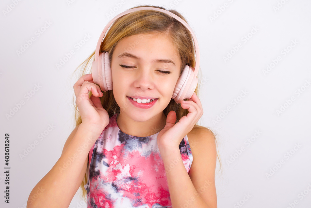 Pleased little caucasian kid girl wearing sport clothing over white background enjoys listening pleasant melody keeps hands on stereo headphones closes eyes. Spending free time with music Pleased little caucasian kid girl wearing sport clothing over white background enjoys listening pleasant melody keeps hands on stereo headphones closes eyes. Spending free time with music