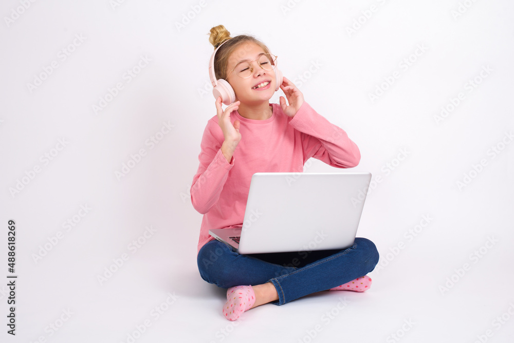 Beautiful caucasian teen girl sitting with laptop in lotus position on white background with headphones on head, listens to music, enjoying favourite song with closed eyes, holding hands on headset. Beautiful caucasian teen girl sitting with laptop in lotus position on white background with headphones on head, listens to music, enjoying favourite song with closed eyes, holding hands on headset.