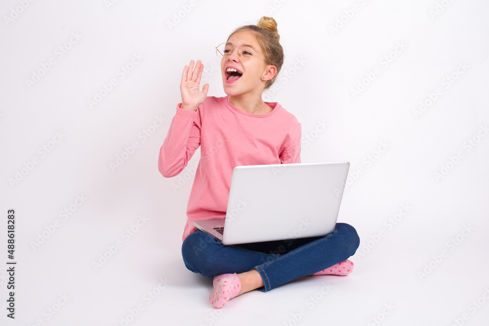 Beautiful caucasian teen girl sitting with laptop in lotus position on white background look empty space holding hand near her face and screaming or calling someone. Beautiful caucasian teen girl sitting with laptop in lotus position on white background look empty space holding hand near her face and screaming or calling someone.