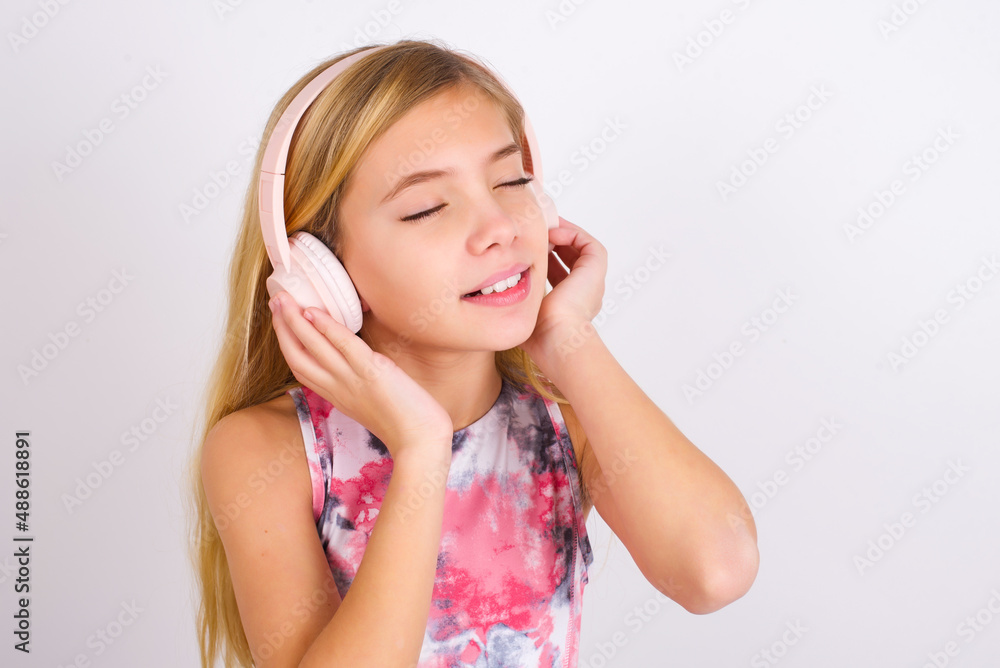 little caucasian kid girl wearing sport clothing over white background with headphones on her head, listens to music, enjoying favourite song with closed eyes, holding hands on headset. little caucasian kid girl wearing sport clothing over white background with headphones on her head, listens to music, enjoying favourite song with closed eyes, holding hands on headset.