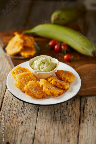 Patacones or tostones, typical Ecuadorian appetizer that consists on fried green plantain slices. It’s accompanied with guacamole and served on a traditional plate with a wooden and rustic background.