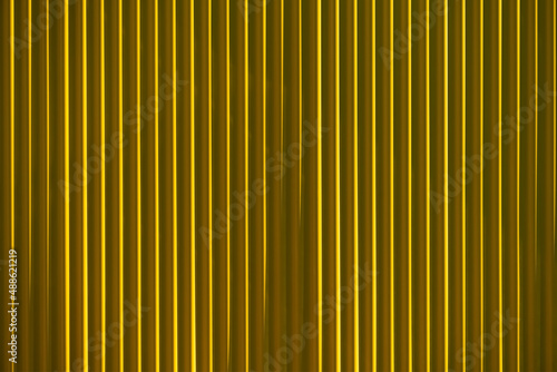 Yellow facade on a modern building wall. Monochrome background of colored profiled sheet metal and illuminated by sunlight from the side of the architecture. Parallel vertical lines, light and shadow.