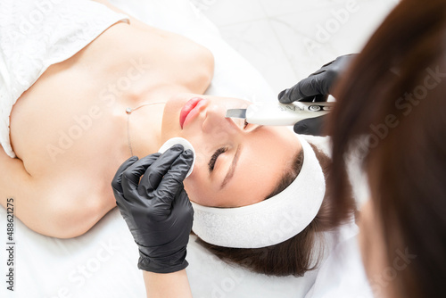 Ultrasound facial peeling. Ultrasonic facial cleansing at cosmetology clinic. Cosmetologist with ultrasonic scrubber works with woman's face skin. Skin cleaning from black dots and other impurities.