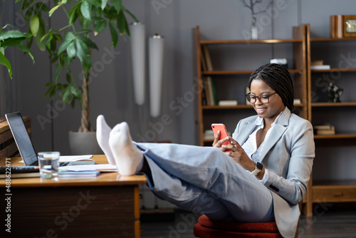 Lazy african american businesswoman procrastinating at workplace sit with feet on desk use smartphone, chat with friend or scroll social media. Entrepreneur female in formalwear rest with mobile phone