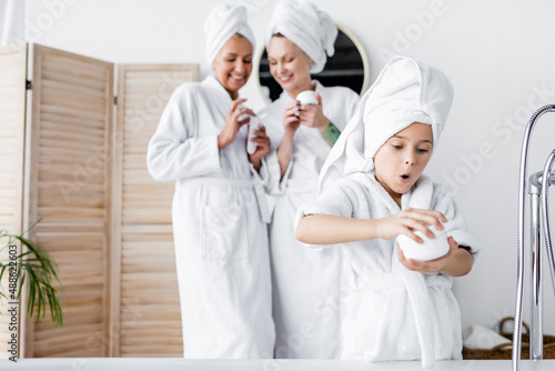 Amazed girl in bathrobe holding cosmetic cream near blurred mothers in bathroom