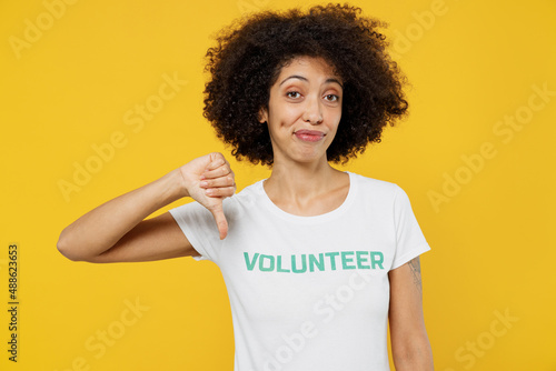 Young woman of African American ethnicity in white volunteer t-shirt showing thumb down dislike gesture isolated on plain yellow background. Voluntary free work assistance help charity grace concept