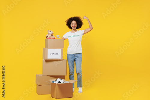 Full body young woman of African American ethnicity in white volunteer t-shirt near boxes with presents show muscles isolated on plain yellow background. Voluntary free work assistance help concept. photo