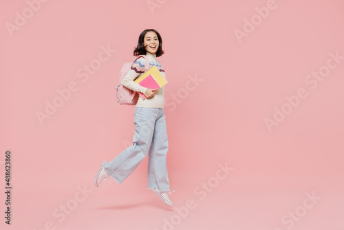 Full size happy excited teen student girl of Asian ethnicity wear sweater hold backpack books jump high look camera isolated on pastel plain pink background Education in university college concept