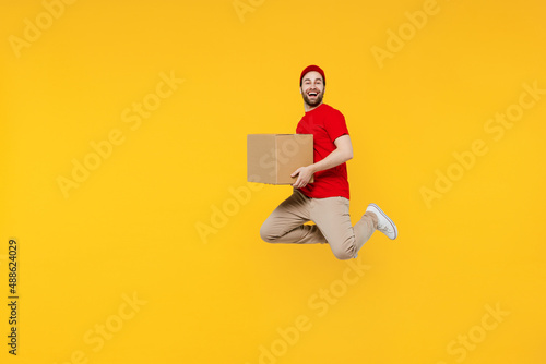Side view full body delivery employee man in red cap T-shirt uniform workwear work as dealer courier jump high hold cardboard box isolated on plain yellow background studio portrait. Service concept photo