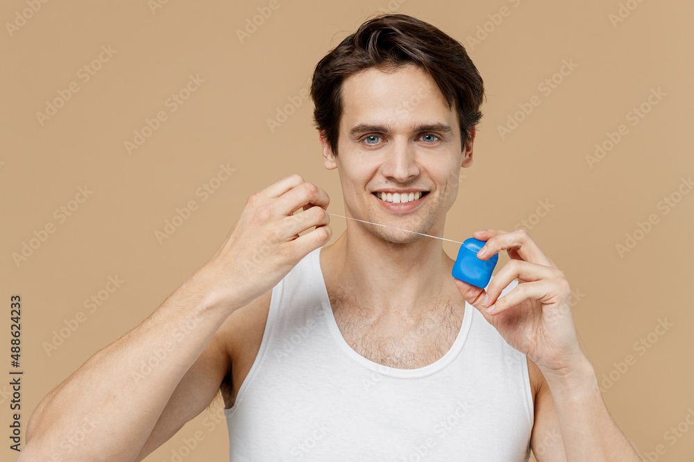 Attractive smiling happy young man 20s perfect skin in undershirt hold using dental floss isolated on pastel pastel beige background studio portrait. Skin care healthcare cosmetic procedures concept. Attractive smiling happy young man 20s perfect skin in undershirt hold using dental floss isolated on pastel pastel beige background studio portrait. Skin care healthcare cosmetic procedures concept.