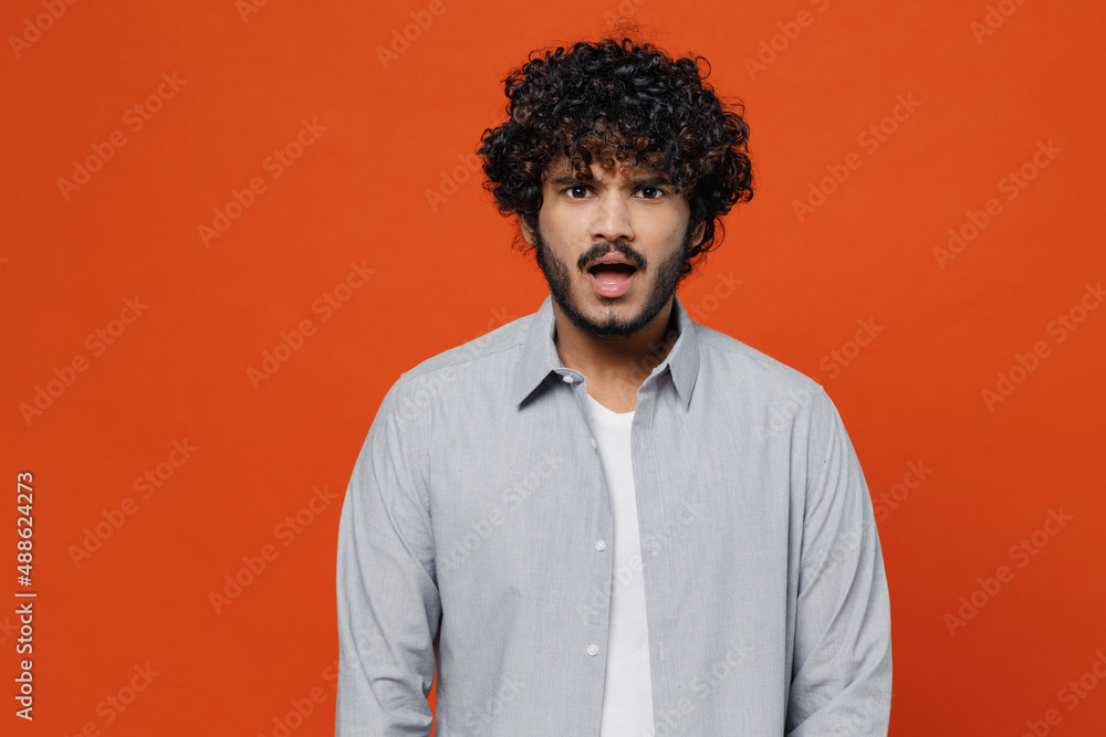 Shocked surprised stupefied displeased sullen amazed young bearded Indian man 20s years old wears blue shirt looking camera keeping mouth wide open isolated on plain orange background studio portrait. Shocked surprised stupefied displeased sullen amazed young bearded Indian man 20s years old wears blue shirt looking camera keeping mouth wide open isolated on plain orange background studio portrait.