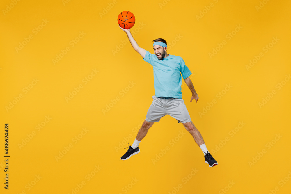 Full length caucasian young fitness trainer instructor sporty man sportsman in headband blue t-shirt hold ball play basketball game jump high isolated on plain yellow background Workout sport concept