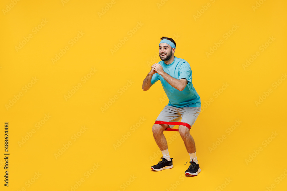 Full body side view happy young fitness trainer instructor sporty man sportsman in headband blue t-shirt use fitness elastic bands do squats isolated on plain yellow background. Workout sport concept.