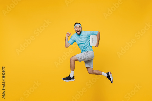 Full size side view strong young fitness trainer instructor sporty man sportsman wear headband blue t-shirt jump high run fast hold scales isolated on plain yellow background Workout sport concept