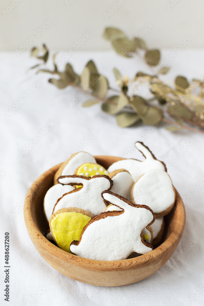 Festive pschal gingerbread in the form of eggs and rabbits with colored icing in a bowl on a white background with willow branches. The concept of traditional holiday food. Vertical orientation. Festive pschal gingerbread in the form of eggs and rabbits with colored icing in a bowl on a white background with willow branches. The concept of traditional holiday food. Vertical orientation.