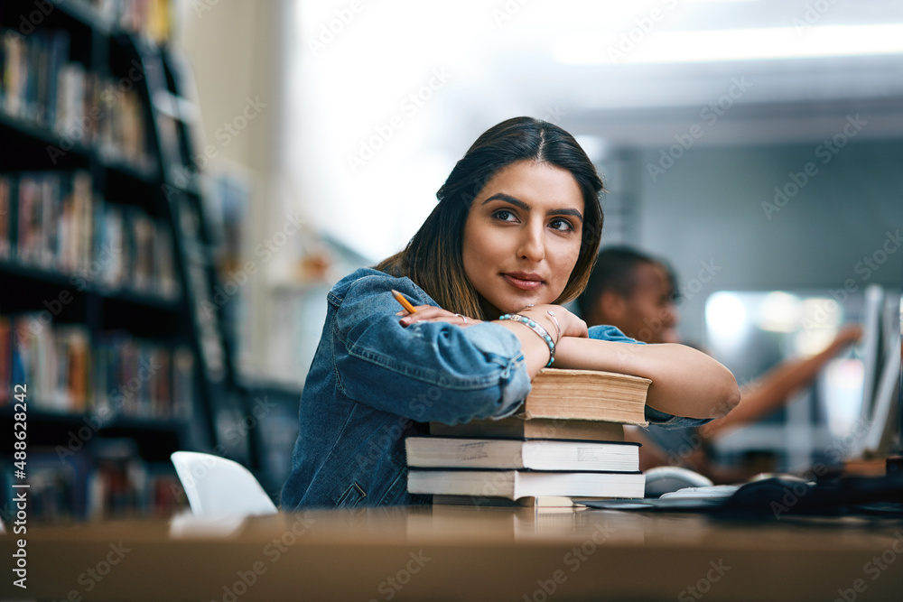 Where dreams are turned into reality. Shot of a young woman resting on a pile of books in a college library and looking thoughtful. - obrazy, fototapety, plakaty 