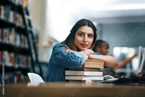 Where dreams are turned into reality. Shot of a young woman resting on a pile of books in a college library and looking thoughtful.