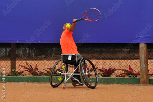 Jovem cadeirante jogando tenis em quadra de saibro com camiseta laranja photo
