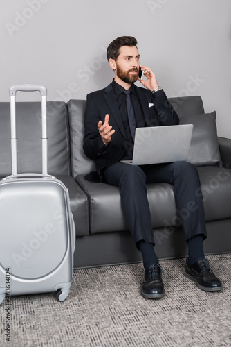 bearded businessman talking on smartphone near laptop and luggage in hotel room