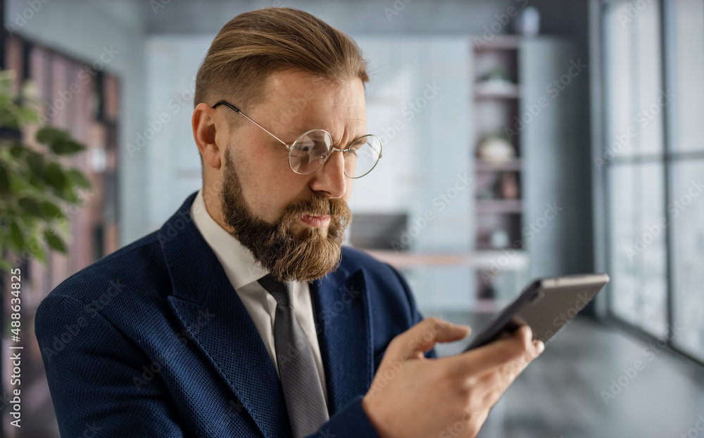 Confident businessman in eyeglasses and stylish suit using modern smartphone while sitting at office. Modern gadget for work and lifestyle. Confident businessman in eyeglasses and stylish suit using modern smartphone while sitting at office. Modern gadget for work and lifestyle.