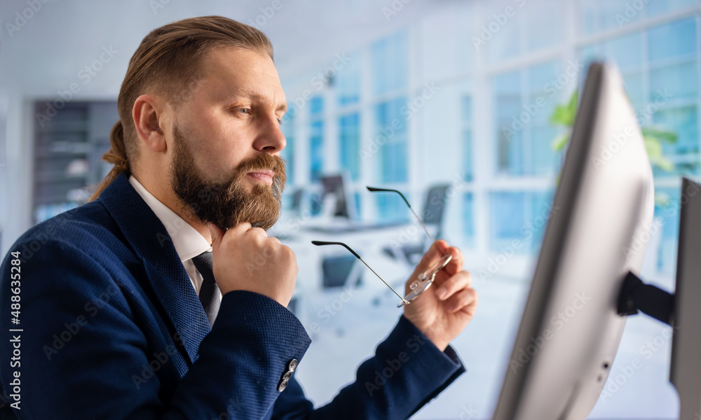 Side view of focused bearded man in stylish suit sitting at modern office and looking on computer monitor. Concept of business, work and technology. Side view of focused bearded man in stylish suit sitting at modern office and looking on computer monitor. Concept of business, work and technology.