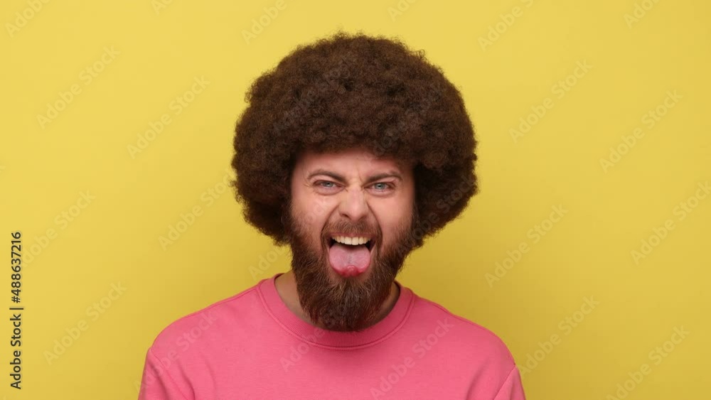 Childish bearded man with Afro hairstyle showing out tongue with naughty disobedient grimace, making face, wearing pink sweatshirt. Indoor studio shot isolated on yellow background. Childish bearded man with Afro hairstyle showing out tongue with naughty disobedient grimace, making face, wearing pink sweatshirt. Indoor studio shot isolated on yellow background.